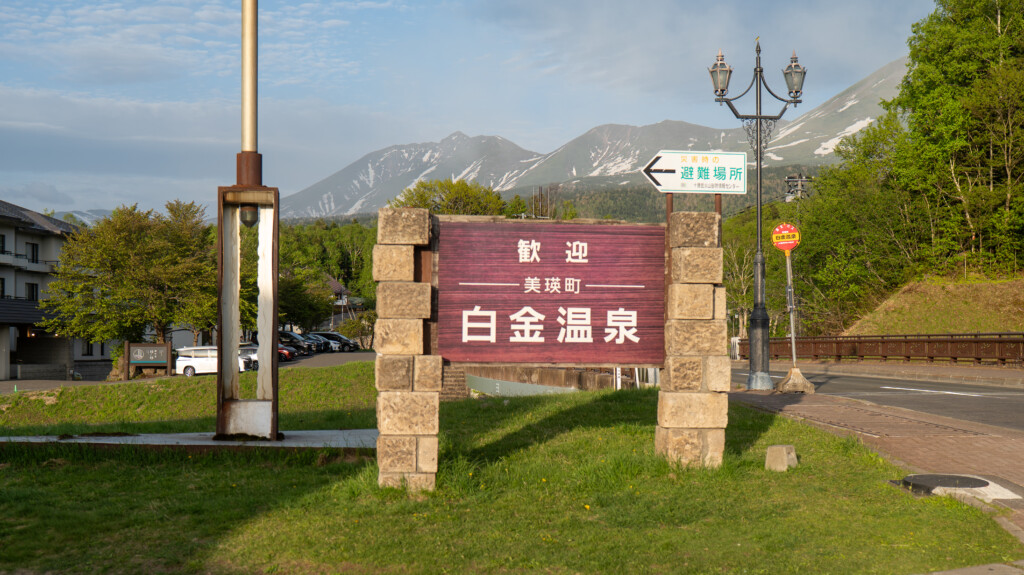 美瑛町白金温泉の入口看板。背後には十勝岳連峰と白金温泉街の風景が広がる